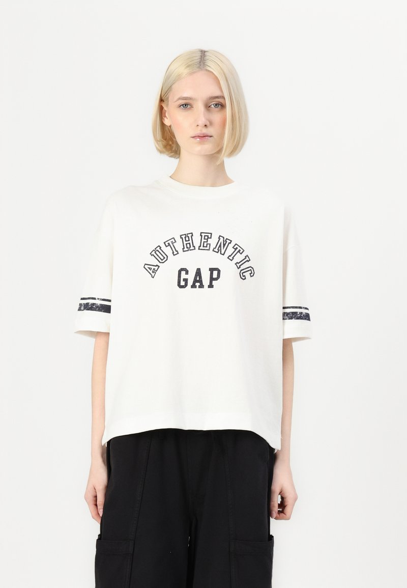 Bílý bavlněný tričko s černým nápisem "AUTHENTIC GAP", zdobené černobílými pruhovanými akcenty na rukávech. Volný střih, kulatý výstřih.