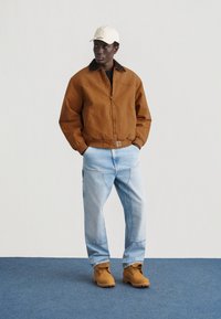 Άνδρας με καφέ μπουφάν, ανοιχτό μπλε τζιν, καφέ εργασιακές μπότες και μπεζ καπέλο του μπέιζμπολ στέκεται σε μπλε χαλί μπροστά σε λευκό τοίχο.