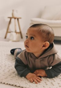 Niemowlę ubrane w dwukolorowy hoodie w odcieniach szaro-beżowym, o miękkiej teksturze. Pełza po kremowym dywanie o fakturze, z rączkami opartymi na powierzchni.