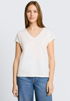Jeune femme aux cheveux lisses châtain clair, portant un t-shirt blanc à manches courtes et col en V et un jean bleu, debout devant un fond uni gris clair.