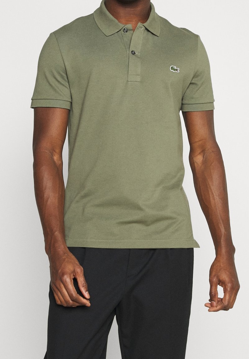 Homme portant un polo vert olive à manches courtes avec deux boutons et un petit logo brodé sur la poitrine, associé à un pantalon noir.