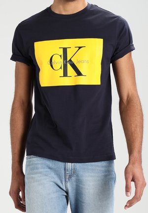 Man draagt een donkerblauw T-shirt met opgerolde mouwen, voorzien van een grote gele rechthoek en een zwart "cK Calvin Klein Jeans" logo, gecombineerd met lichtblauwe spijkerbroek.