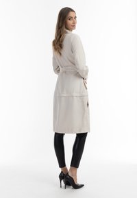 Trench coat bej deschis, cu talie accentuată printr-un brâu, având un design curat și cusături subtile. Purtați peste colanți din piele negri și pantofi cu toc.