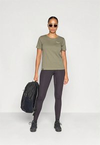 Alyvuogių žalios spalvos trumpomis rankovėmis marškinėliai su balta logotipu, derinami su juodomis tamprėmis. Modelis laiko juodą kuprinę ir dėvi akinius nuo saulės.