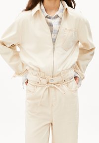 Beige zipjacka med krage; elastiska byxor med knytning i midjan. Mjuk tyg, kontrasterande sömmar och bröstfickdetalj.