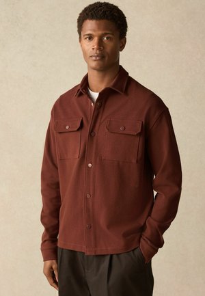 Brown button-up shirt with two chest pockets, made of textured fabric. Features a collar and a relaxed fit, paired with dark trousers.