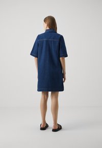 Robe en denim bleu foncé, à manches courtes, coupe droite, col, avec des détails de couture visibles. Associée à des sandales noires.