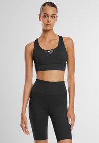 Sort sports-bh og høj taljeret shorts sæt i strækbart stof, der har en bred kant og logo på bh'en. Glat tekstur og tætsiddende design.