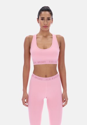 Kobieta ubrana w dopasowany, jasnoróżowy krótki top i legginsy Ted Baker Sport, stojąca z ramionami luźno opuszczonymi, zwrócona przodem, na jednolitym tle.