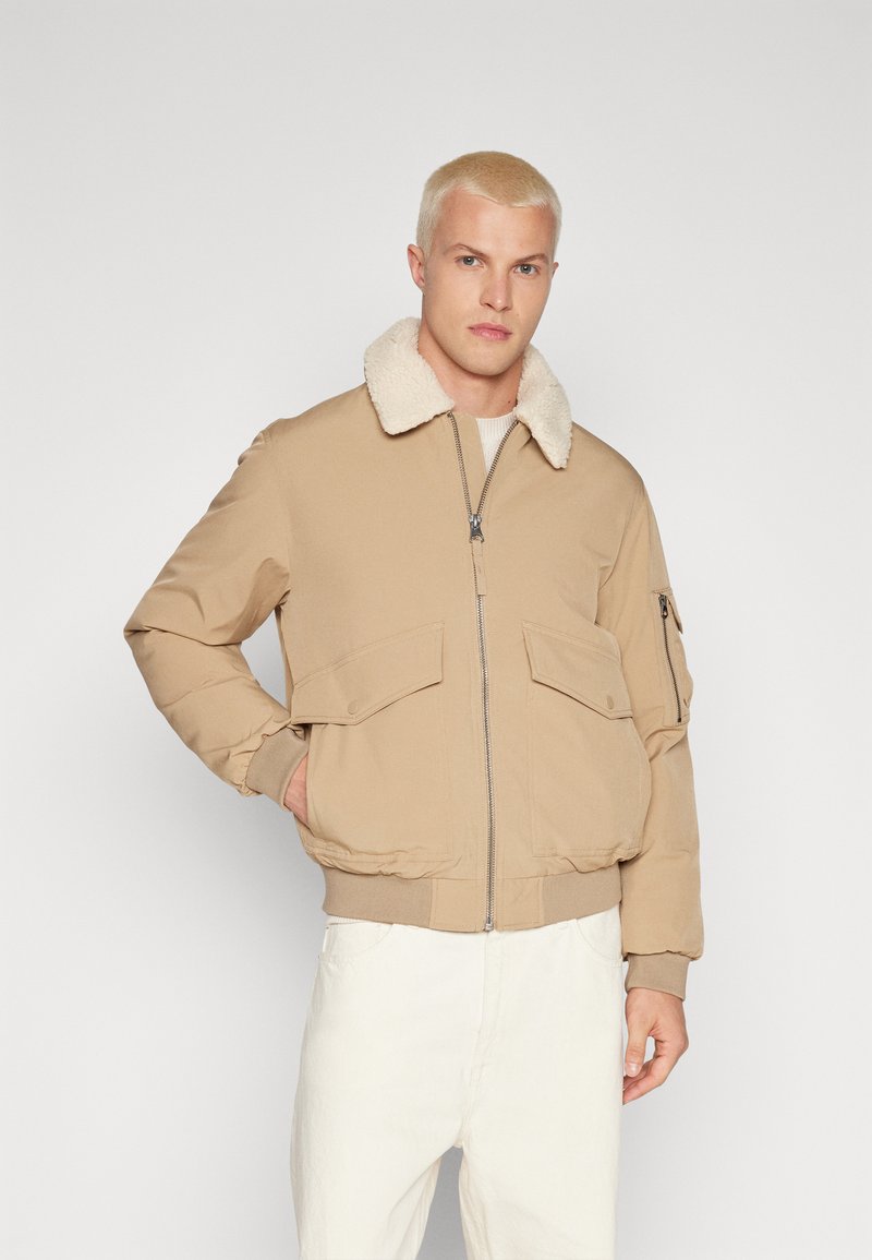 Chaqueta bomber beige con cuello de borreguillo, cierre de cremallera y dos bolsillos frontales. Textura suave con puños y dobladillo acanalados.