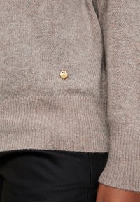 Pull en maille beige avec un ourlet et des poignets côtelés, orné d'un petit logo doré près du bas sur le côté. Texture lisse.