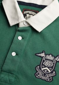 Polo vert avec col et boutons blancs, arborant un écusson sombre avec le texte "Superdry 1954 The Kings of Quality" sur la poitrine.