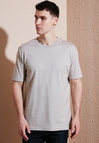 Světle šedé tričko s krátkým rukávem vyrobené z hladkého materiálu, s jemně reliéfním logem na přední straně a klasickým kulatým výstřihem.