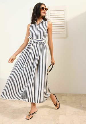 Naine valges varrukateta kleidis, millel on vertikaalsed tumesinised triibud, päikeseprillid, mustad kontsakingad, hoides väikest musta käekotti, seisab siseruumis.