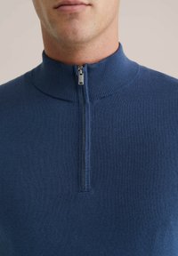 Pulover tricotat bleumarin cu guler înalt și închidere cu fermoar pe jumătate. Textură netedă cu accente striate la guler și zona fermoarului.