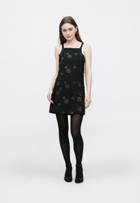 Juoda suknelė su tekstūruota medžiaga, puošta gėlių raštu. Rankovių neturinti, aptempta forma, derinama su juodomis pėdkelnėmis ir batais.