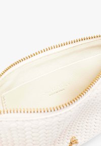 Bolsita de cuero blanco texturizado con cremallera dorada, que presenta un sutil logo en el interior y un pequeño acento de calavera en la parte frontal.