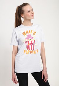 Hvid t-shirt med et farverigt grafisk motiv af en bjørn i en stribet popcornspand, med teksten "HVAD'S POPPING?" i lys gul og orange.