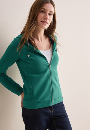 Felpa verde con zip, texture a coste, fodera interna bianca, due tasche laterali e design aderente, abbinata a jeans scuri.