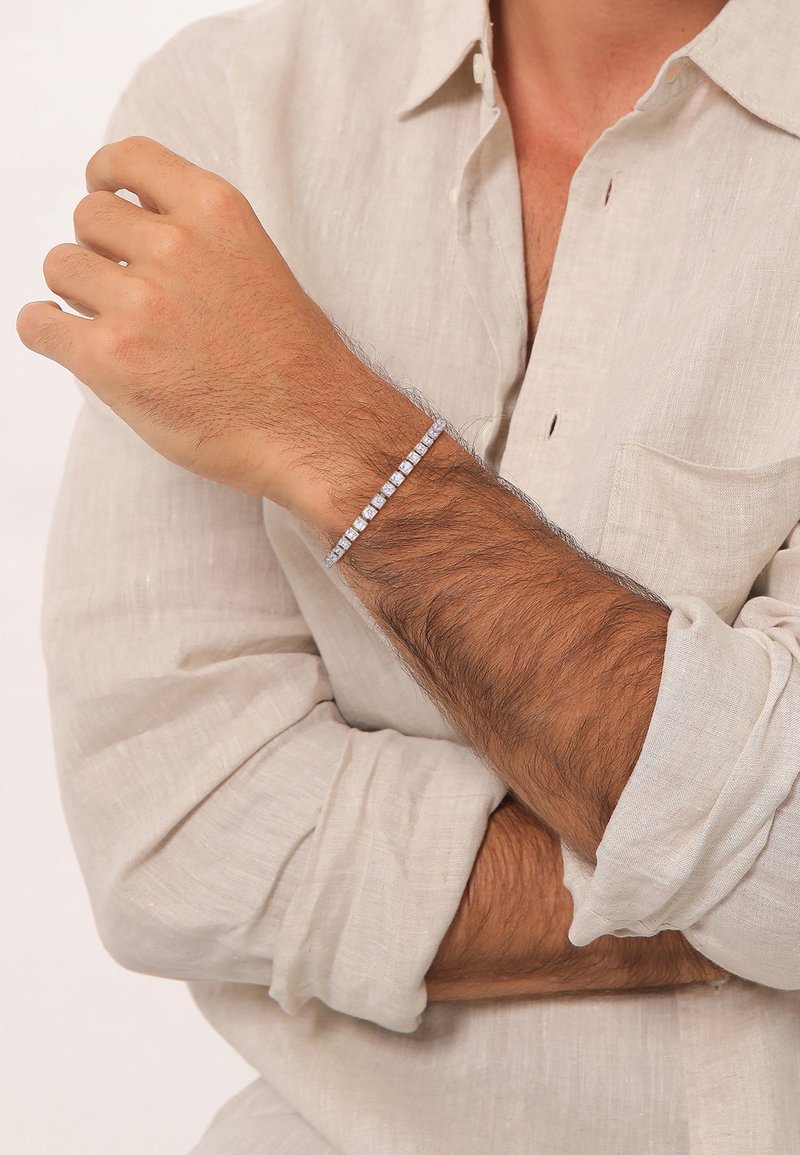 Hõbedane käekett ruudukujuliste aksessuaaridega, peene ahela küljes, kantud randmel, millel on heledat pruuni juukseid. Kanga särk on beež.