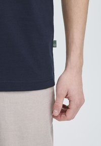 Donkerblauwe katoenen t-shirt met korte mouwen, voorzien van een klein groen merklabel aan de zijkant, gecombineerd met lichtbeige broek.