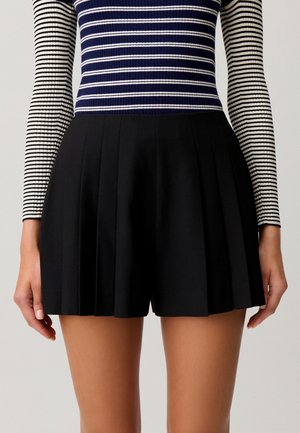 Mujer que lleva pantalones cortos negros plisados y una camiseta de manga larga con rayas horizontales azul marino y blancas, vista desde la cintura hasta la mitad del muslo.