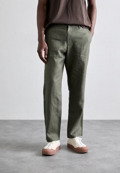 Green linen trousers with a relaxed fit, paired with white sneakers featuring a pink rubber sole. Simple design, no visible patterns.