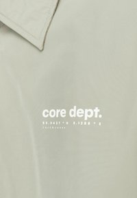 Gaiši pelēka jaka ar lielu balto drukāto logo "core dept." ar ģeogrāfiskajām koordinātēm, marķēta ar Jack & Jones. Gluda tekstūra.