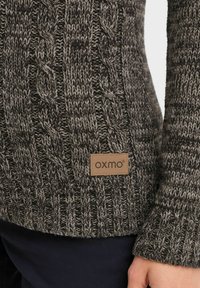 Kabelstrikket genser i mørkegrå og svart. Har ribbestrikkede mansjetter og et lærmerke med logo. Teksturert, tykk strikkemønster.