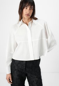 White, cropped button-up shirt with pointed collar and long sleeves, featuring a textured fabric and subtle seam detailing. Paired with black floral pants.