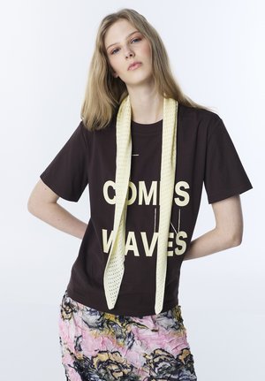 Młoda kobieta o długich blond włosach, ubrana w ciemną koszulkę z dużym napisem, teksturowany żółty szalik i kolorową, kwiecistą spódnicę.