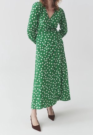Kobieta ubrana w zieloną sukienkę kopertową z długim rękawem w białe kropki oraz brązowe buty z noskiem w szpic, stojąca na jednolitym tle.