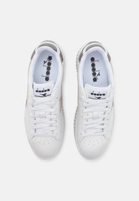Baskets blanches avec des accents en cuir texturé beige et gris. Elles présentent un design à lacets, une zone de perforation au niveau des orteils et un logo sur la languette.