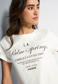 Hvid t-shirt med korte ærmer, der har mørkebrune tekster: "L.A. Palm Springs," "Embrace Connection," og "Studio Society." Glat bomuldsstof.