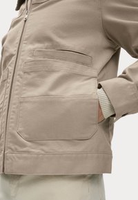 Giacca beige con grandi tasche frontali, chiusura con zip e una mano parzialmente dentro la tasca destra, indossata sopra un maglione con polsini a costine.