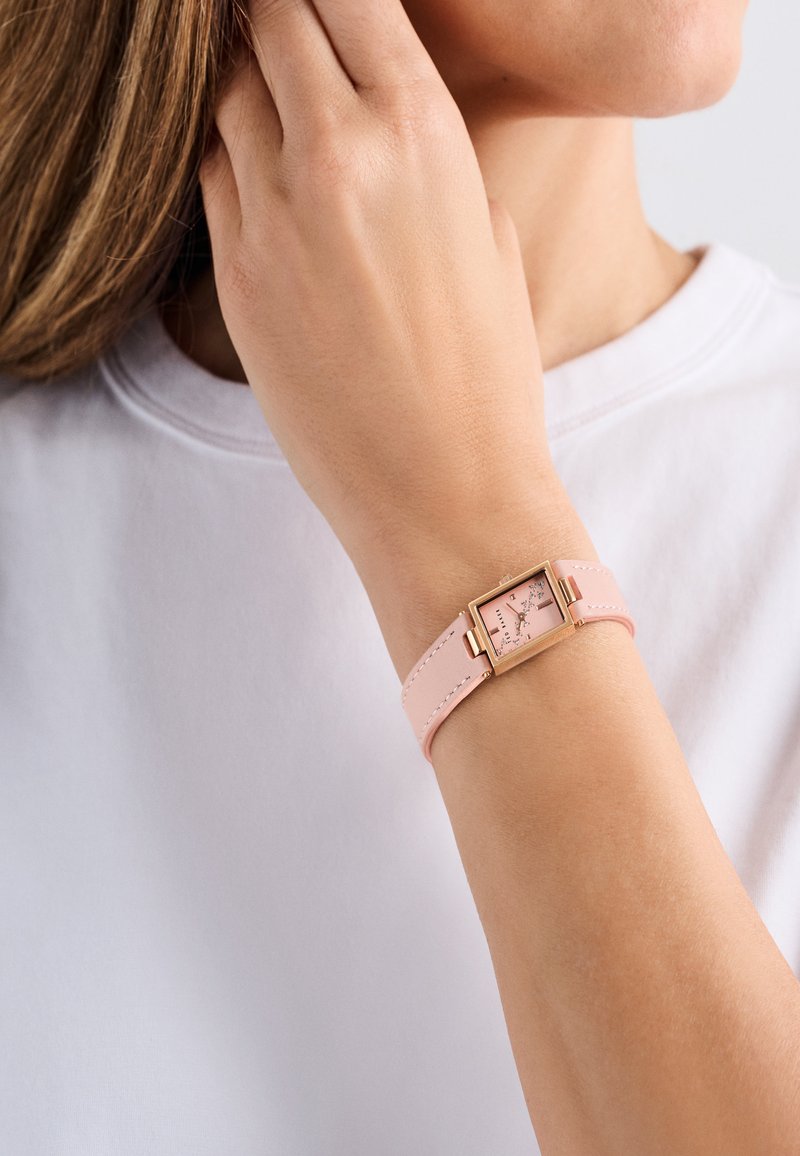 Roze leren strapwatch met een rechthoekige gouden metalen kast en een lichtroze wijzerplaat met zilveren uuraanduidingen.