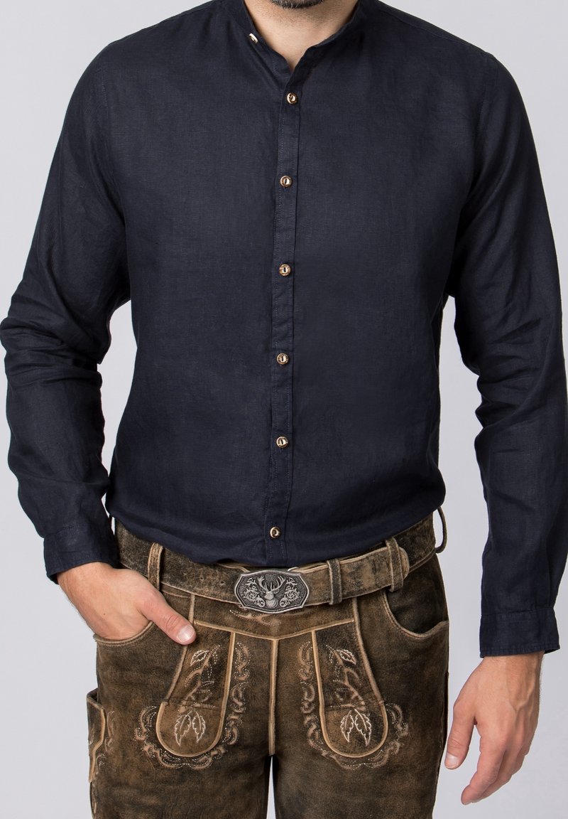 Homme portant une chemise bleu foncé boutonnée rentrée dans un lederhosen en cuir marron brodé avec une boucle de ceinture décorative.