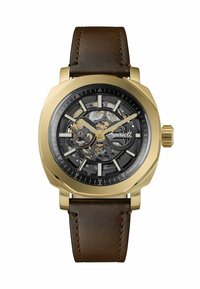 Orologio da polso con quadrante scheletrato color oro, ingranaggi visibili, indicatori neri e dorati, e cinturino in pelle marrone.