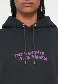 Fekete kapucnis pulóver, amelyen egy lila grafikai szöveges dizájn található, ami így olvasható: "Pase lo que pase nunca para nada," fém végű húzózsinórokkal.