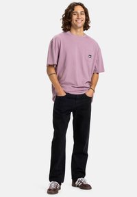 Camiseta corta de manga en color lila con un bolsillo en el pecho y logo, combinada con unos vaqueros rectos negros y zapatillas marrones con detalles en blanco.