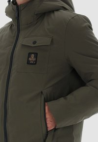 Giacca imbottita verde oliva con cappuccio, dotata di chiusura con zip, tasca sul petto e tasche laterali. Texture liscia con una finitura resistente.