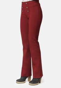 Mujer vestida con pantalones de campana rojos de cintura alta con botones decorativos y botines negros de tacón, de pie contra un fondo neutro.