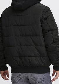 Czarna pikowana kurtka z kapturem, z poziomymi przeszyciami, ściągaczami przy mankietach i jednolitym czarnym kolorem. Materiał wydaje się gładki i lekki.