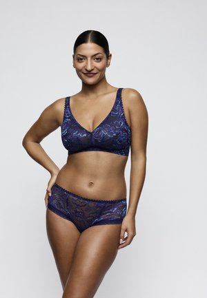 Woman standing and smiling, wearing matching blue and purple floral lace bra and underwear against a plain light background.