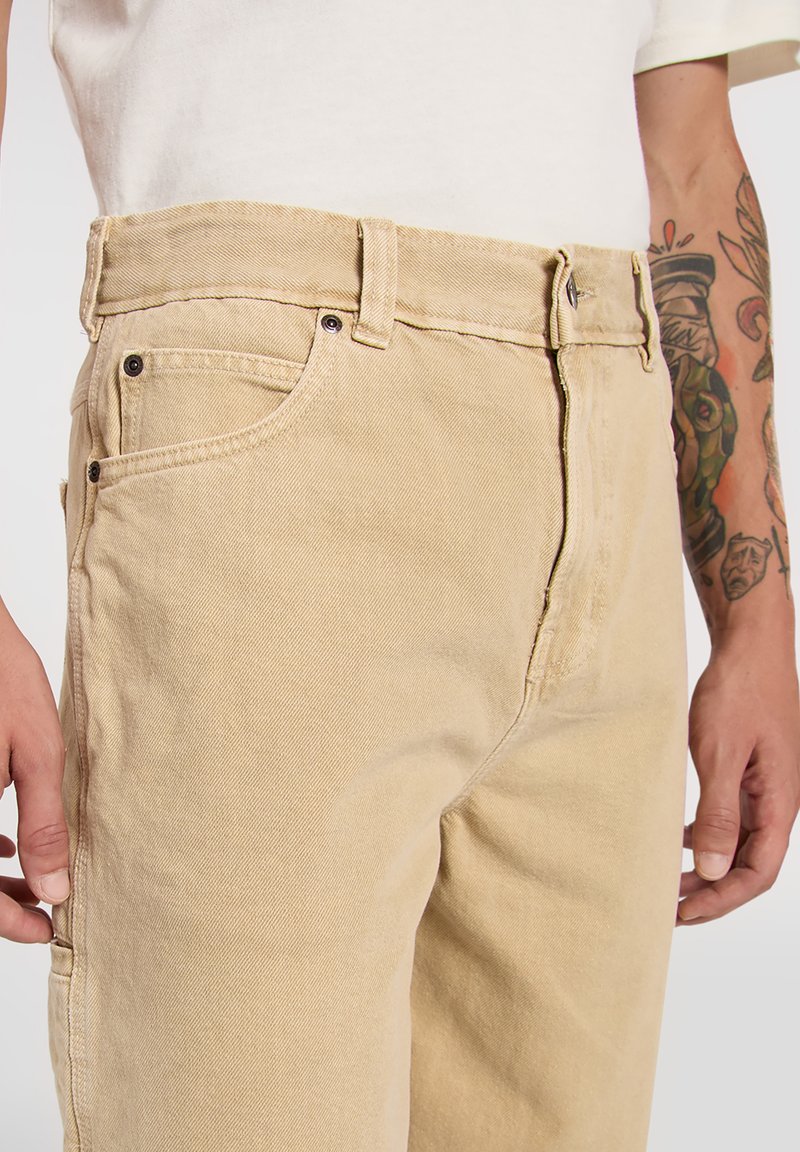 Pantalones de mezclilla beige claro con diseño de pierna recta, que cuentan con cinco bolsillos, cierre de cremallera y detalles de costuras visibles.