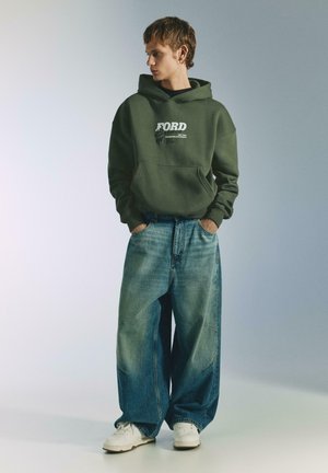 Groene hoodie met "FORD" opschrift, grote zak en geribbelde manchetten, gecombineerd met oversized vervaagde blauwe spijkerbroek en witte sneakers.