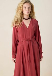 Femme blonde aux longs cheveux ondulés, portant une robe longue à manches longues de couleur rouge profond, avec une taille nouée et un collier choker en perles sombres.