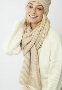Écharpe beige en tricot avec un design texturé à côtes et un accent replié, portée avec un bonnet assorti et un pull crème.