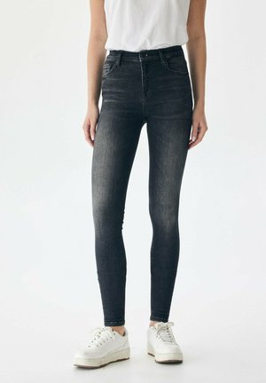 High-waisted black skinny jeans made of denim, featuring a faded texture, five pockets, and a zipper fly, paired with white sneakers.