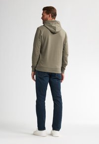 Sudadera verde oliva con capucha ajustable, puños y dobladillo acanalados, combinada con jeans azul oscuro y zapatillas blancas. Diseño simple y limpio.